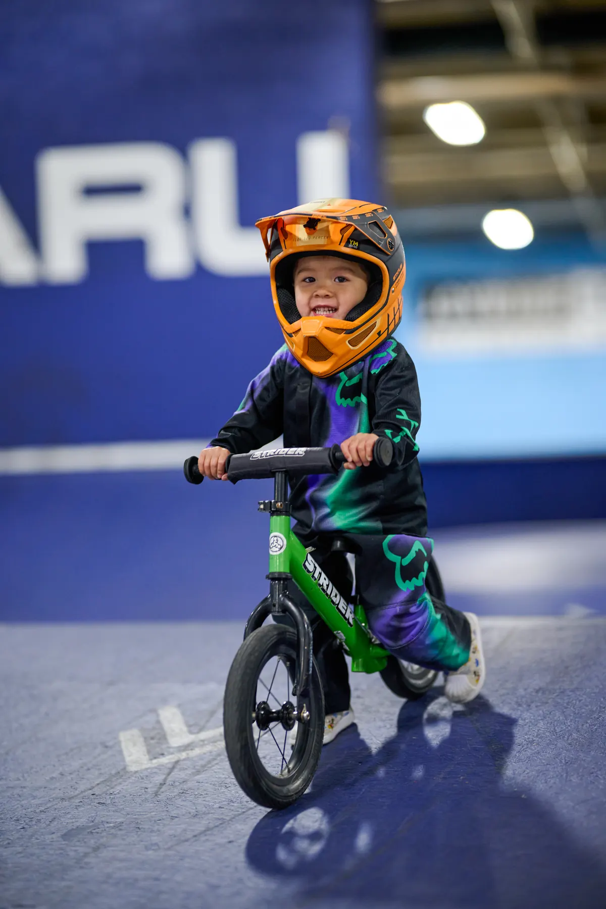 A young child in a full-face helmet smiling while riding a green balance bike at an indoor XKIDS practice park.