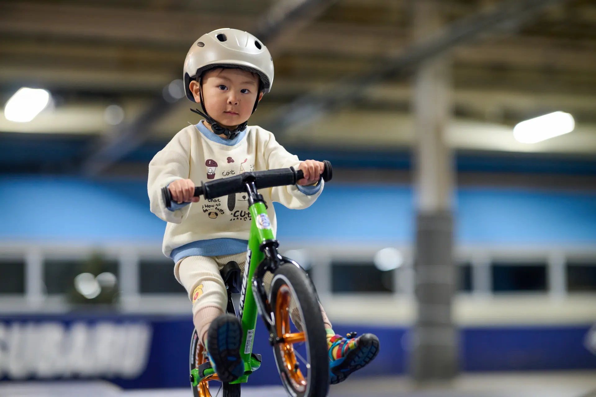 A young child on a green balance bike riding through an indoor XKIDS practice course.