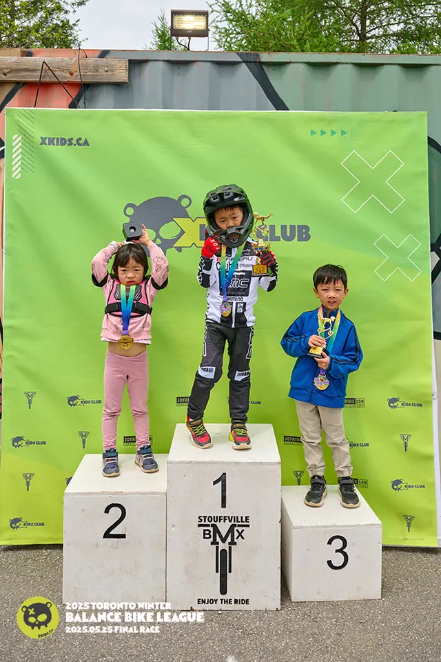 三位 XKIDS 平衡车联赛总决赛者站在 Stouffville BMX 领奖台上,胸前奖牌、手中奖杯,背景是绿色 XKIDS Club 横幅,2025 年 5 月。