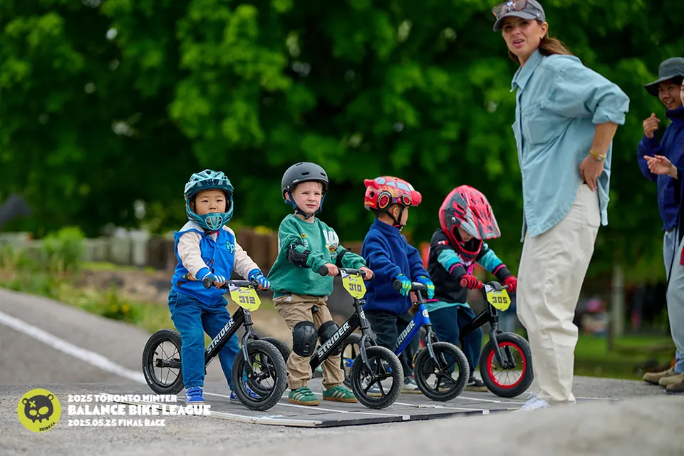 四位 XKIDS 平衡车联赛小骑手在 Stouffville BMX 户外赛道上比赛,戴全盔的教练紧随其后,2025 年 5 月总决赛。