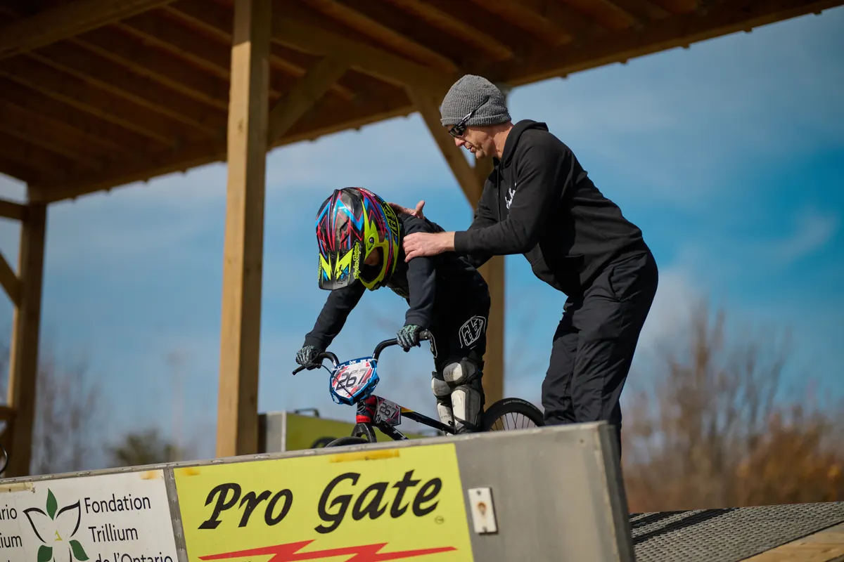 XKIDS 教练在 BMX 起跑门旁检查一位小骑手的装备准备。