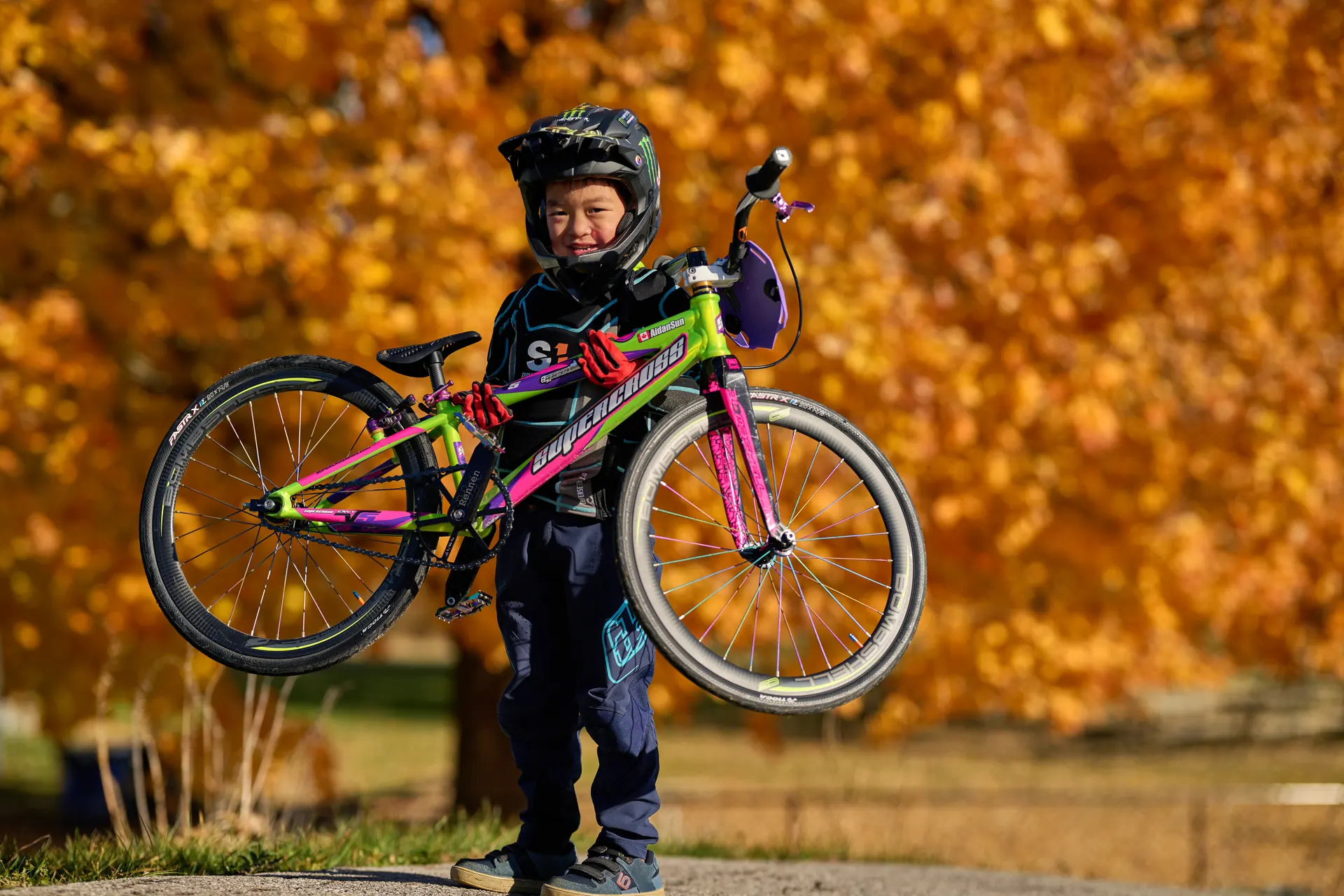 A young XKIDS BMX rider on a colourful bike against autumn track surroundings.