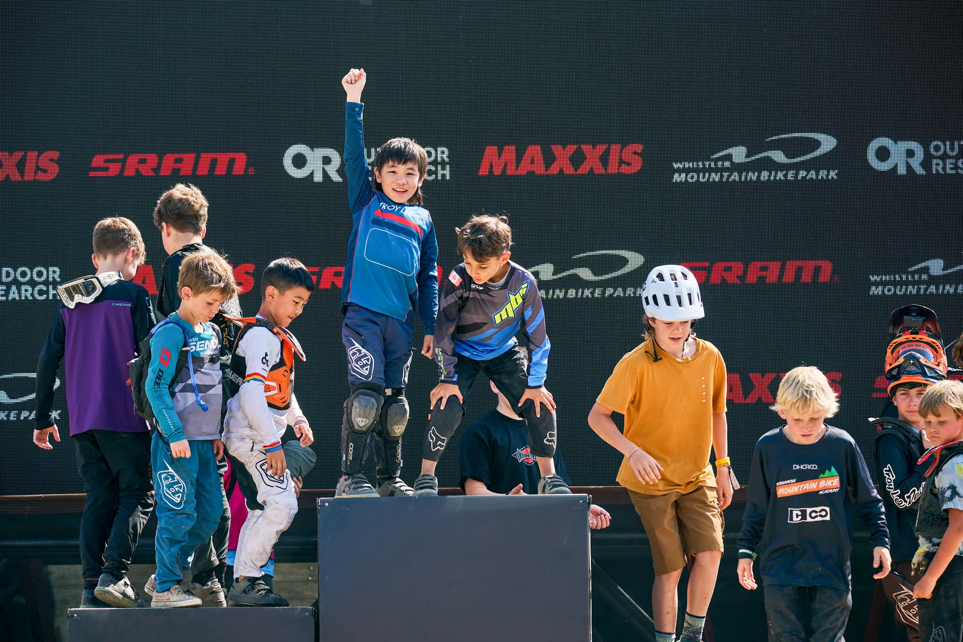 Young XKIDS riders gathered at a mountain bike venue with team jerseys and sponsor banners.