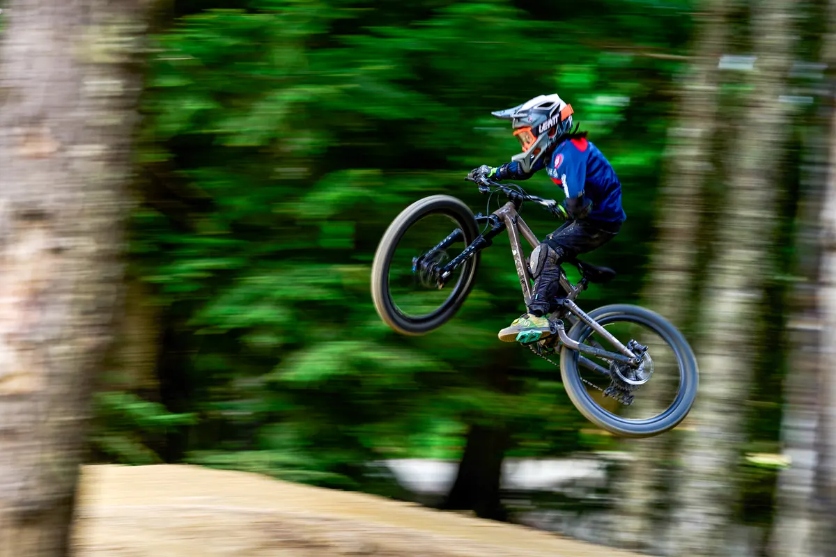 A young XKIDS mountain bike rider mid-jump on a forest trail.