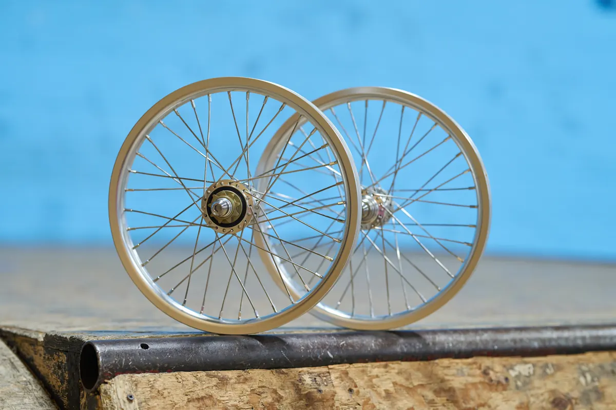 A pair of gold-anodized BMX race wheels with silver spokes resting on a bench against a blue wall.