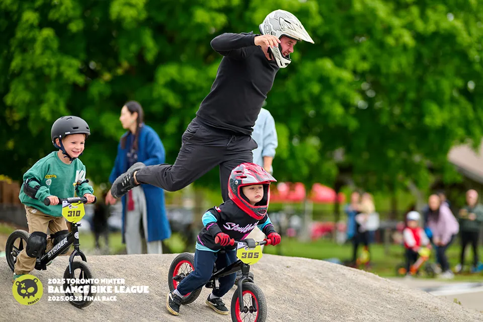 XKIDS 平衡车联赛的两位小骑手在 Strider 平衡车上 — 一位戴粉色头盔,一位穿绿色卫衣 — 在 2025 年 5 月 Stouffville BMX 户外总决赛的 pump 道上较劲,身后是腾跃的教练。